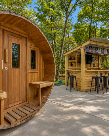 Wooden barrel sauna and outdoor bar structure surrounded by tall trees in a wooded setting