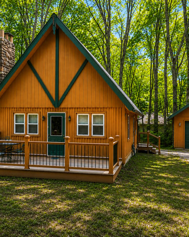 Wooden cabin with green trim and stone chimney surrounded by tall trees, front deck with railing, and detached garage.