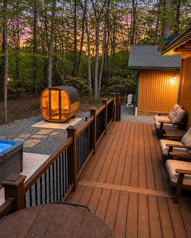 Log cabin deck at dusk with hot tub, sauna pod, Adirondack chairs, and forest backdrop at sunset.
