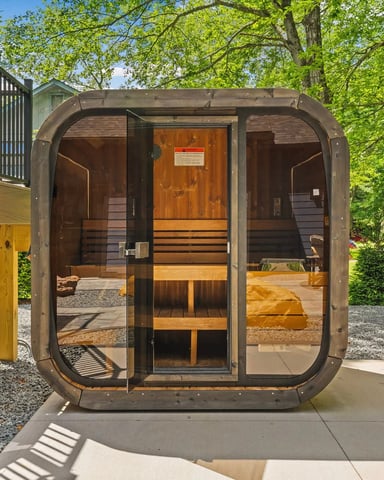 Modern barrel sauna with glass door and wooden interior, surrounded by tall trees and green lawn