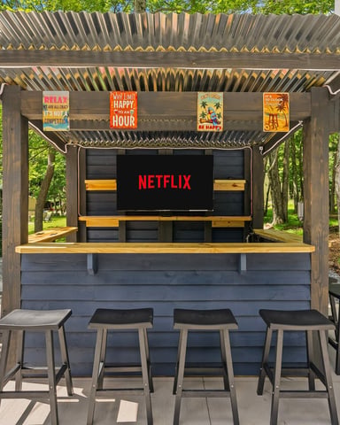 Outdoor Netflix-themed bar structure with slatted pergola roof, bar seating with four stools, and colorful motivational signs in wooded setting.