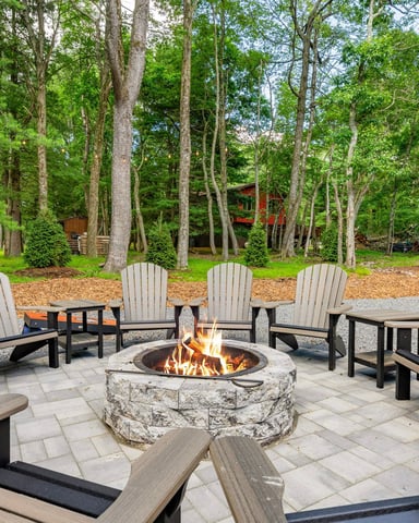 Outdoor fire pit patio with gray Adirondack chairs, stone fireplace, and tall trees surrounding wooded property