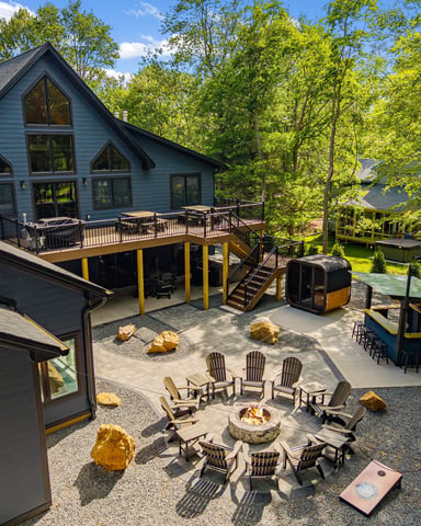 Modern blue house with elevated deck, fire pit surrounded by Adirondack chairs, and wooded grounds with guest structures.