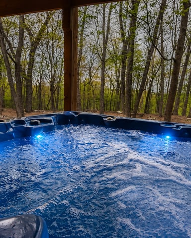 Blue hot tub with LED lights on covered deck overlooking bare trees and green forest