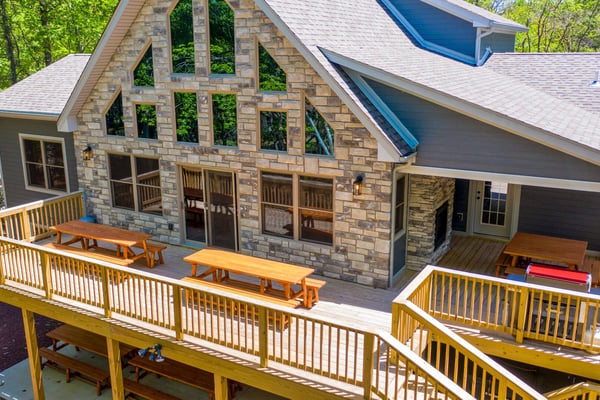 Modern house with stone and siding exterior, wooden deck, and fire pit surrounded by picnic tables and tall pine trees.