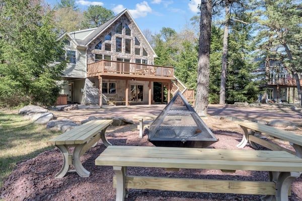 Modern house with stone and siding exterior, wooden deck, and fire pit surrounded by picnic tables and tall pine trees.