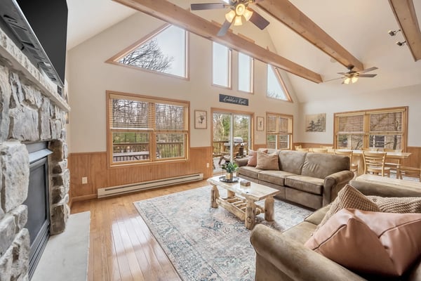 Vaulted living room with stone fireplace, exposed wood beams, multiple windows, ceiling fans, and upholstered seating overlooking wooded landscape.