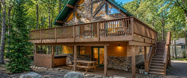 Modern A-frame cabin with stone and wood exterior, wraparound deck, and large windows surrounded by tall trees.