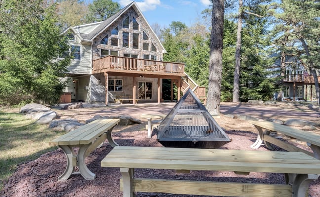 Modern house with stone and siding exterior, wooden deck, and fire pit surrounded by picnic tables and tall pine trees.