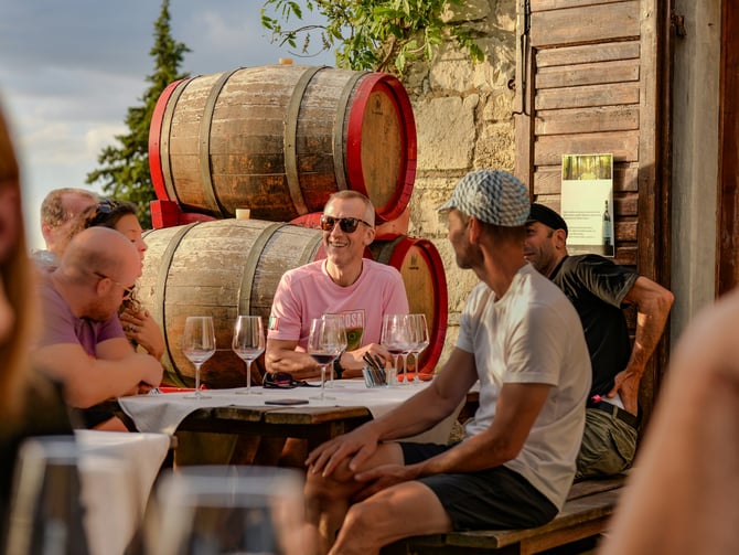 Friends tasting wine at an outdoor table beside oak barrels at a winery