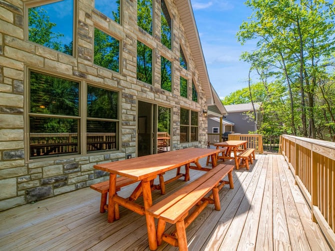 Wooden deck with picnic tables attached to stone house with large windows, surrounded by trees