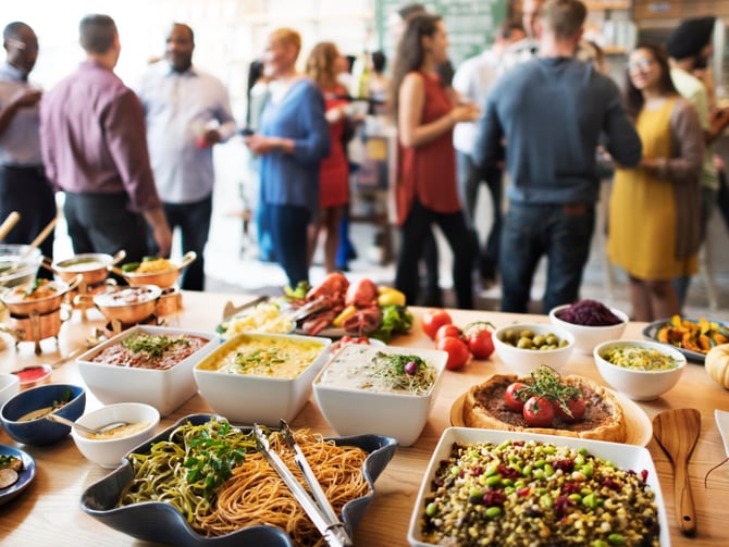 Buffet table with various dishes and fresh vegetables, guests mingling in background at indoor event