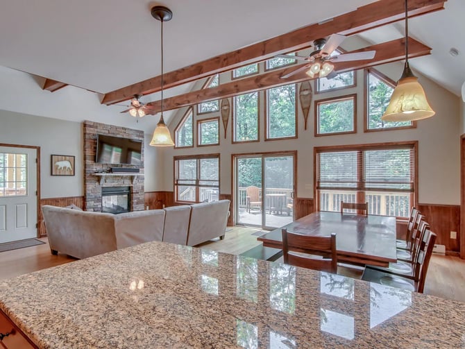 Open-plan living room with granite kitchen island, exposed wood beams, fireplace, and large windows overlooking deck