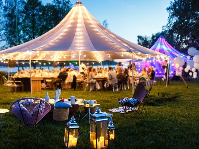 Illuminated circus tent with dining tables and guests at dusk, surrounded by colorful lounge seating and lanterns on grass