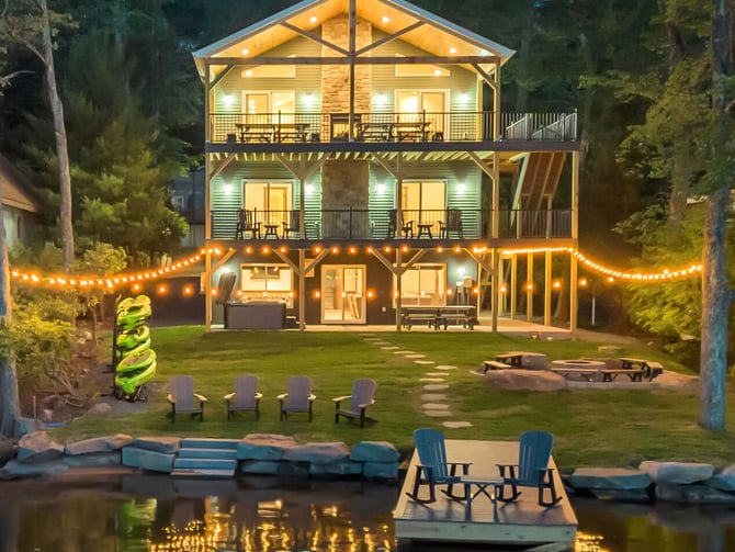 Illuminated three-story waterfront house at dusk with deck, fire pit, string lights, and Adirondack chairs on lawn and dock.