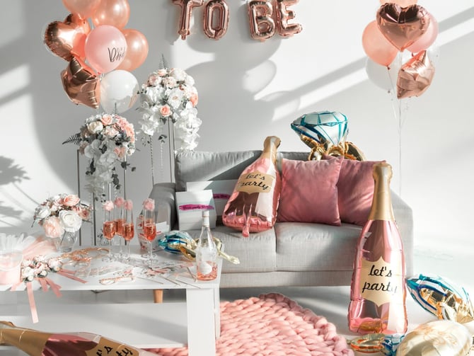 Bride-to-be party setup with rose gold balloons, floral arrangements, champagne bottles, and decorative cushions on gray sofa