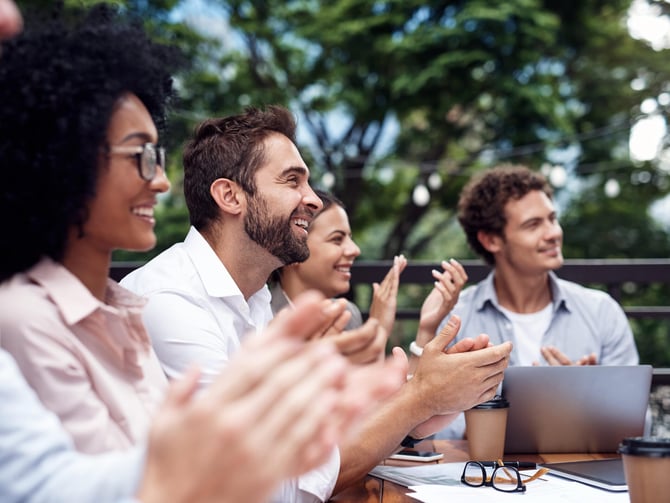 Group of four people clapping outdoors at a table with laptop and papers, trees in background