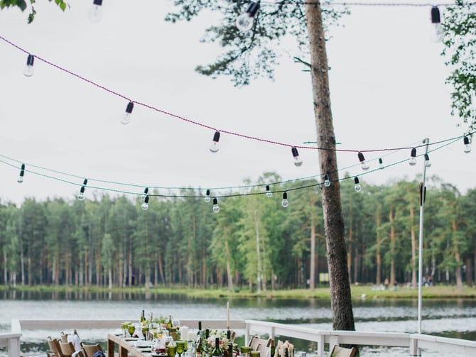 Outdoor lakeside dining setup with long wooden table, string lights, and forest backdrop