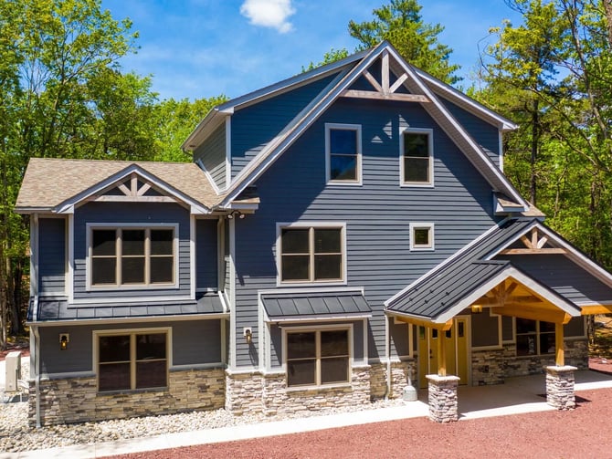 Modern two-story house with dark blue siding, stone foundation, attached garage, and covered porch surrounded by tall trees.