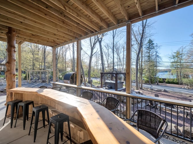 Covered deck with wooden bar counter, black stools, and railing overlooking wooded property with lake view