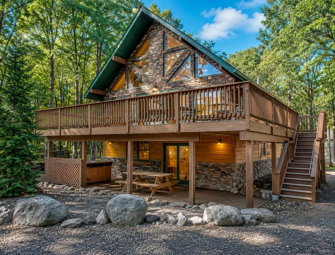 Modern A-frame cabin with stone and wood exterior, wraparound deck, and large windows surrounded by tall trees.